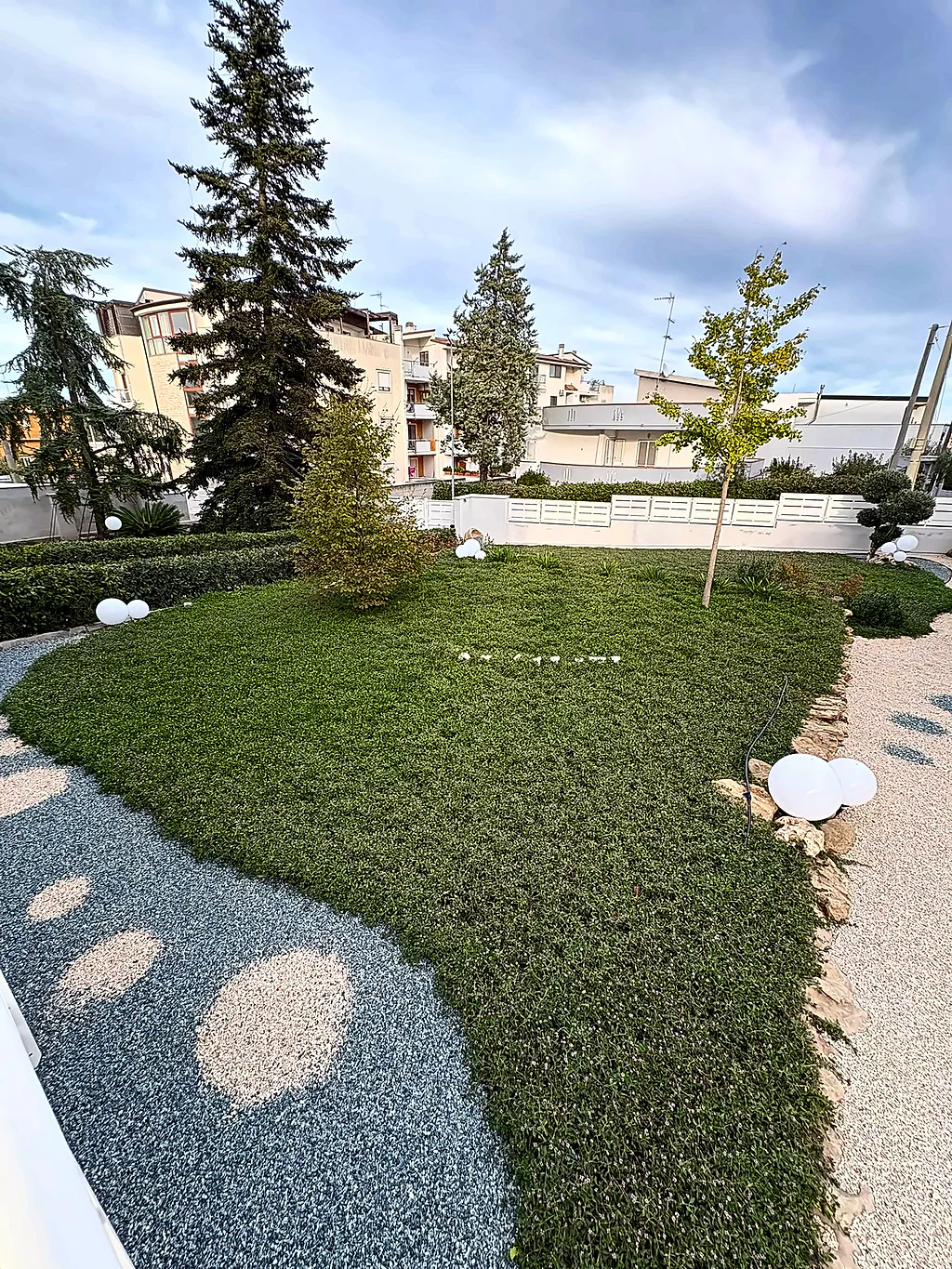 Il prato attraversato da un sentiero di pietre tonde, con un ginkgo sulla destra e altri alberi lungo il bordo