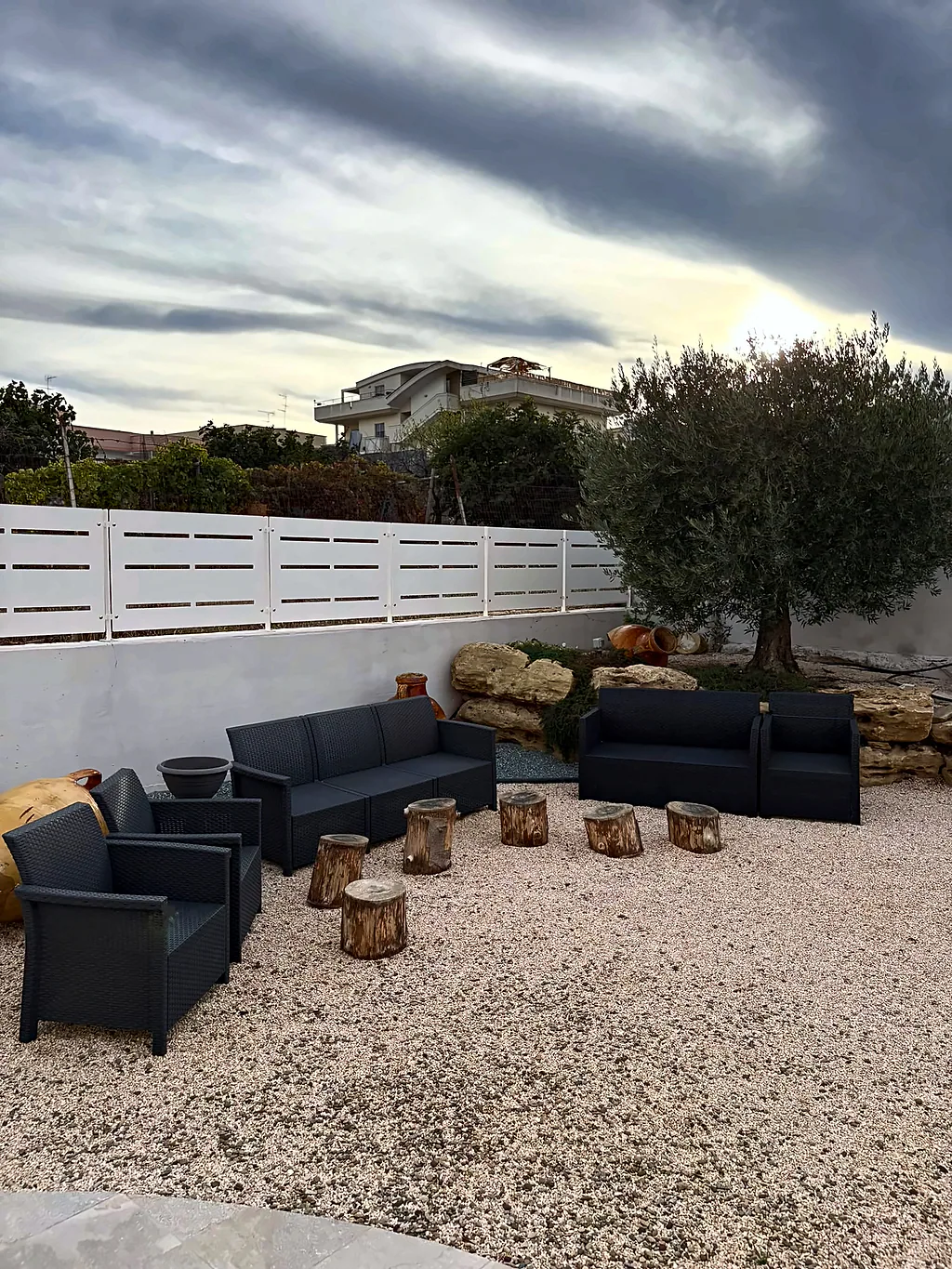 Area relax all'aperto su ghiaia, con divani scuri, sgabelli in tronchi di legno e un ulivo