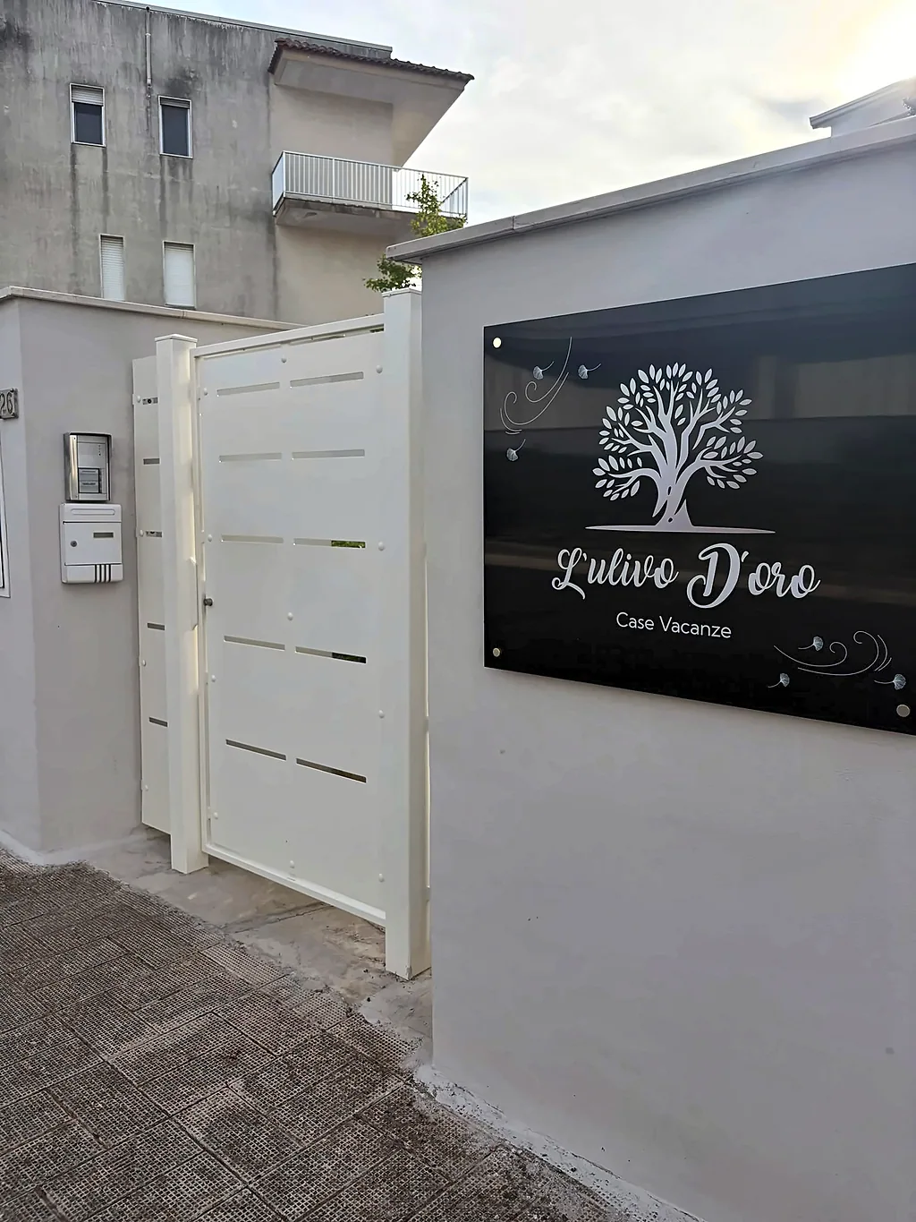 Il cancello d'ingresso bianco con la targa nera «L'Ulivo D'oro – Case Vacanze» e il logo dell'albero
