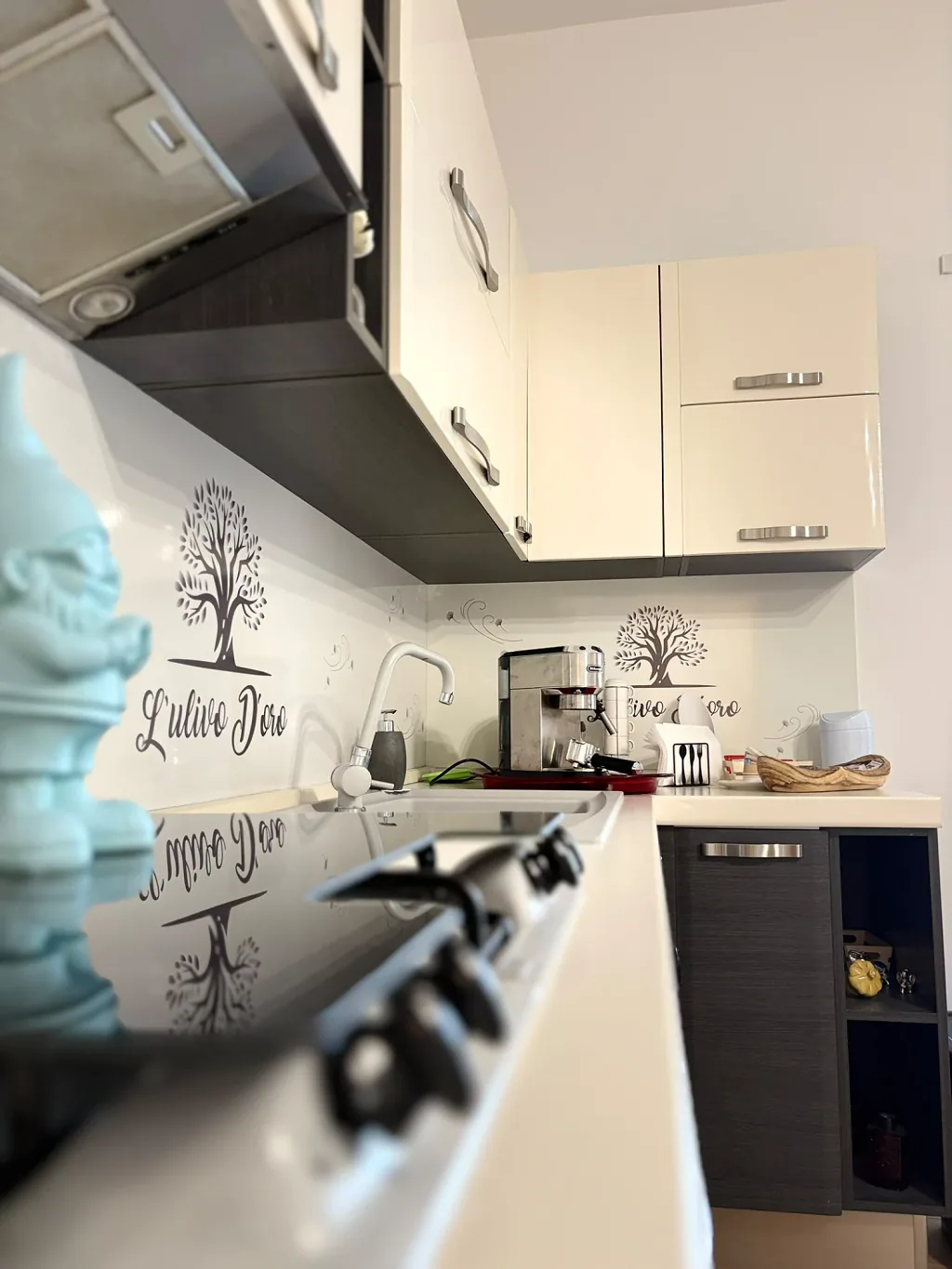 Angolo della cucina con pensili color crema, lavello, macchina per l'espresso e il logo di L'Ulivo D'oro sul paraschizzi; in primo piano, sfocati, il piano cottura e una statuina azzurra